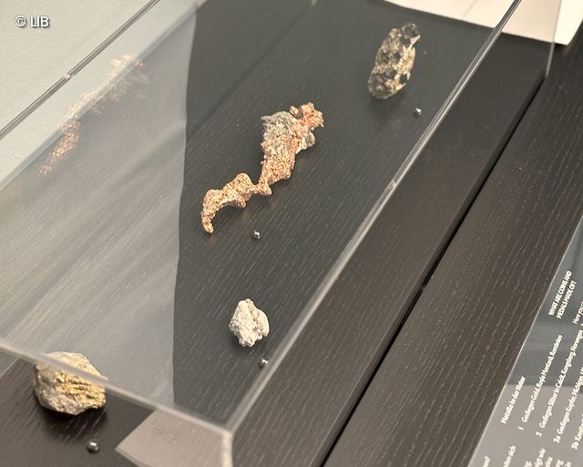 Display case with several irregularly shaped silver and copper ore samples on a dark surface under glass cover
