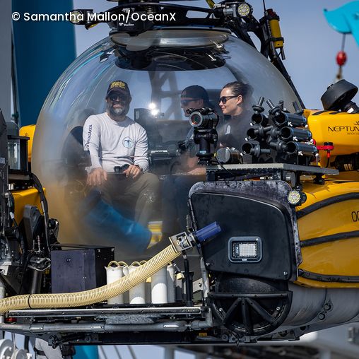 Drei Personen sitzen in einer gelben Tauchkapsel mit kugelförmiger Frontscheibe und technischen Geräten außen am Fahrzeug