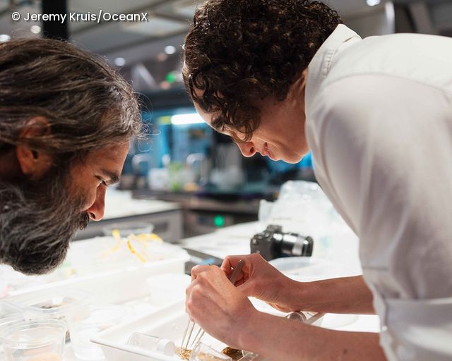 Two people leaning over a table with lab equipment and a camera in the background
