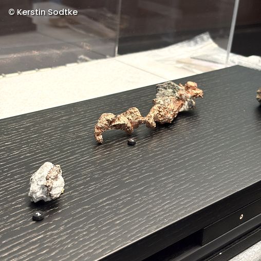 Drei Erze, darunter ein längliches kupferfarbenes Erz in der Mitte, auf schwarzer Holzoberfläche mit kleinen schwarzen Nummernschildern.