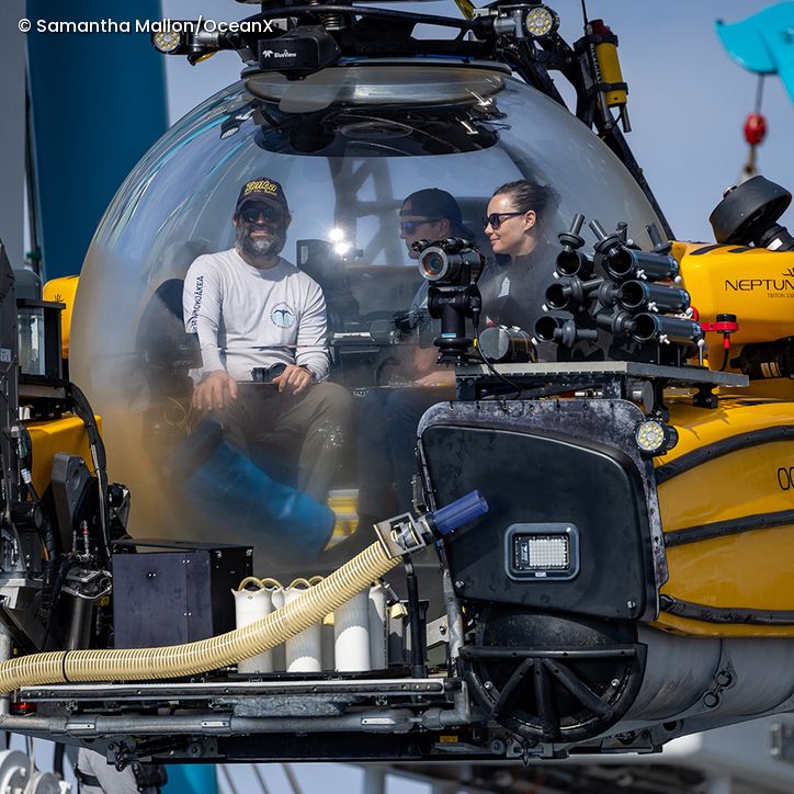 Drei Personen sitzen in einer gelben Tauchkapsel mit kugelförmiger Frontscheibe und technischen Geräten außen am Fahrzeug